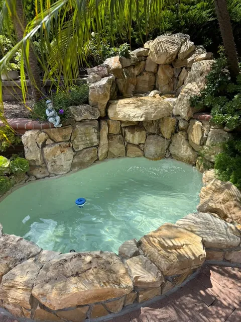 Stacked rock waterfall fountain with the basin refilled and water running, framed by palms and garden plants
